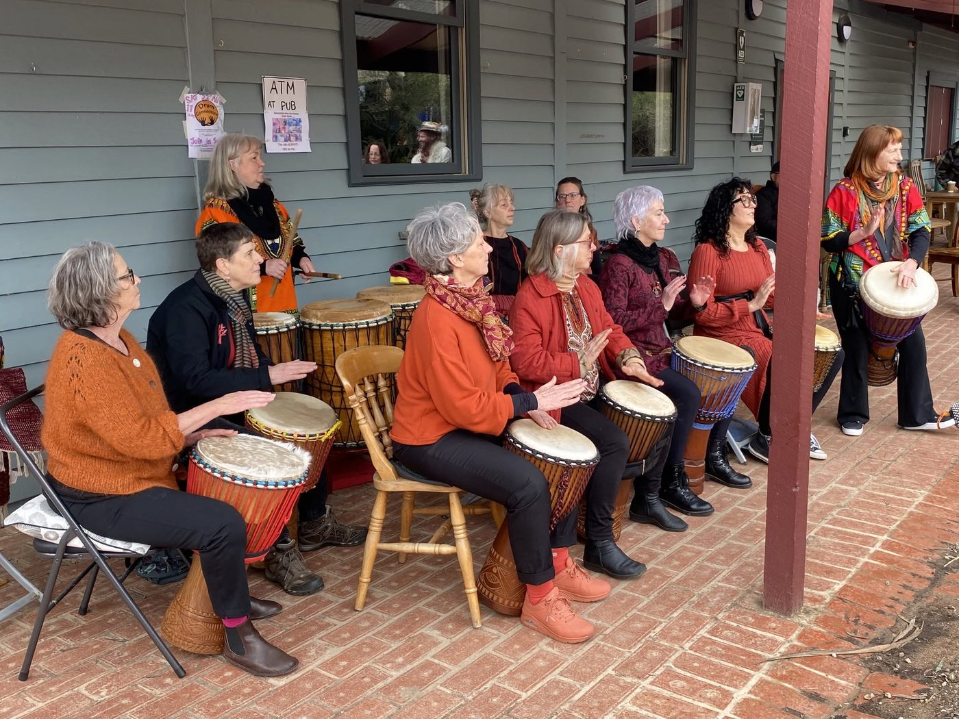 A group of drummers seated in a row on a brick verandah, playing djembes together