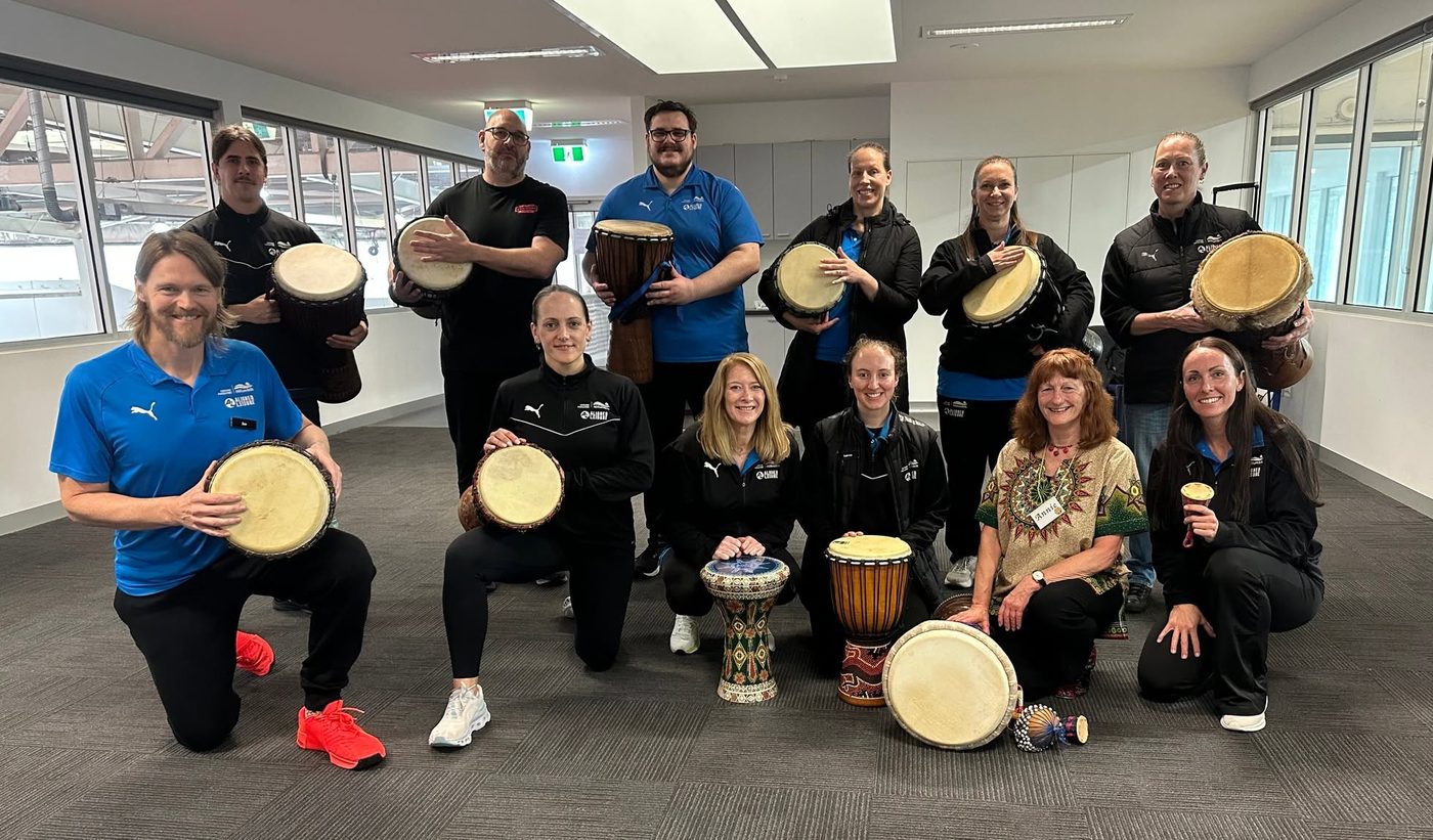 A smiling workplace team holding djembes after a drumming workshop with Annie